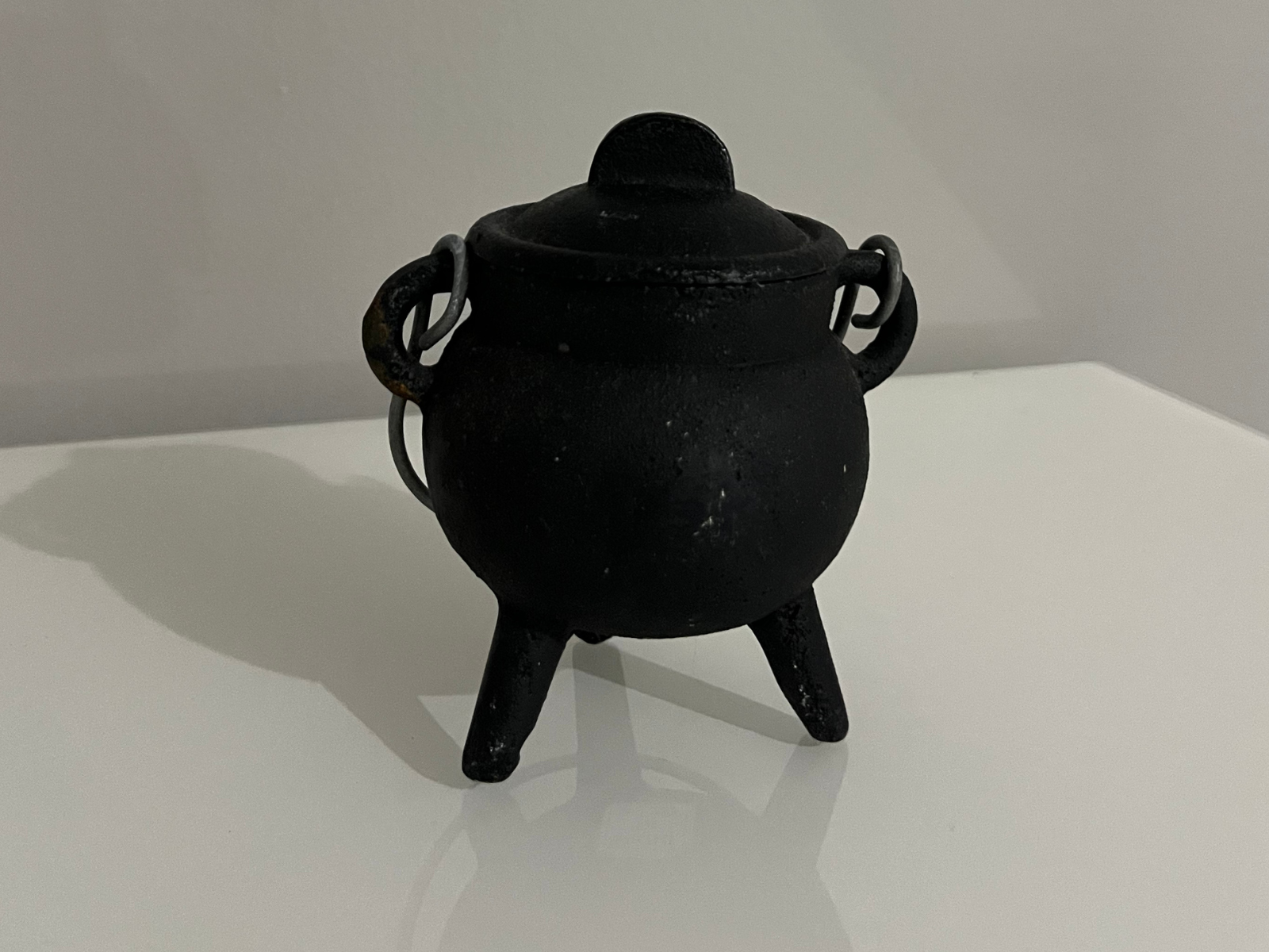 This is a photo of a small black cauldron, taken from the front at eye level. It shows the round body on three short legs, with two side handles and a lid on top. The matte surface has small textures and imperfections that make it look worn and real. The light creates a soft shadow under the cauldron, which shows its shape and volume. This angle is useful as a reference because it shows the proportions and the silhouette clearly.