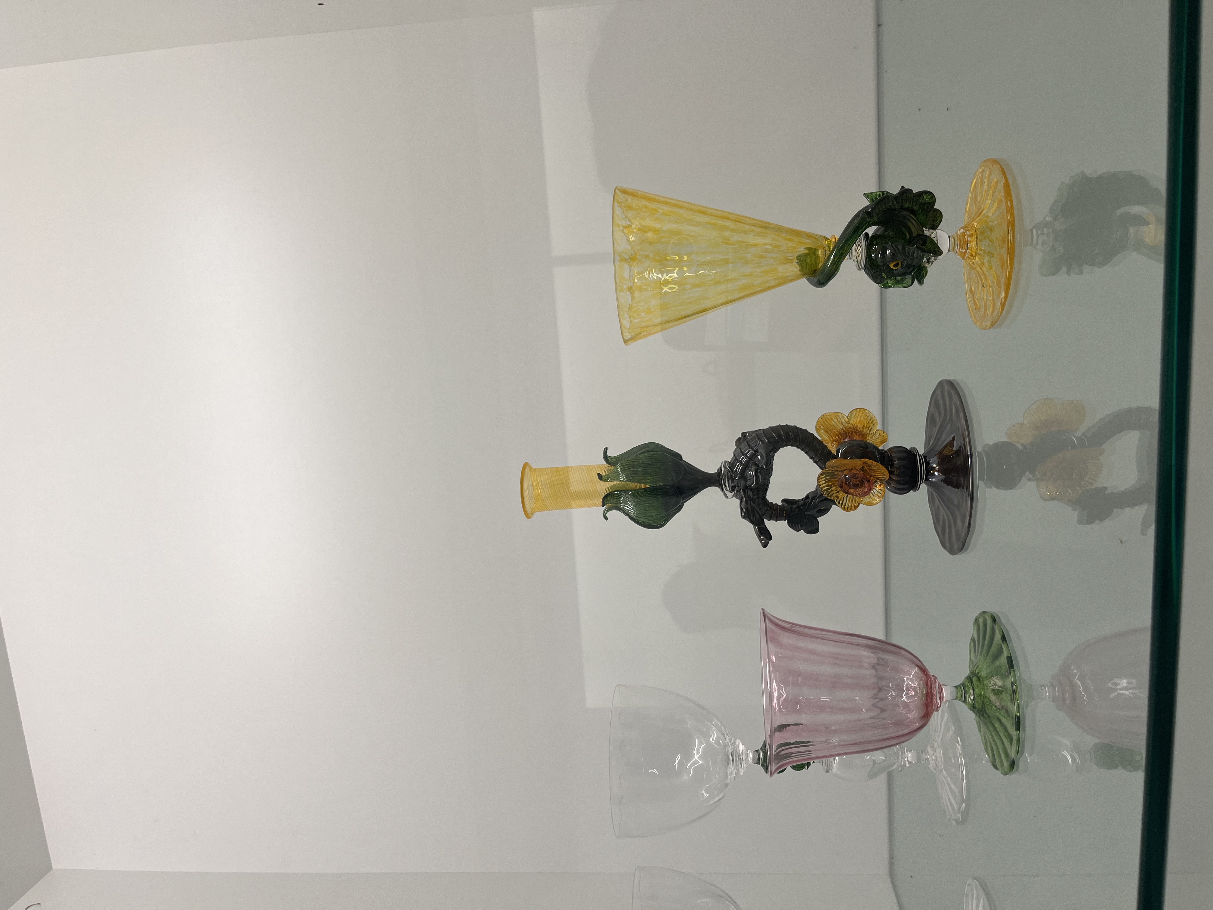 The material is colored glass that has been hand-blown and shaped into delicate cup forms. It is rigid, because glass is hard and cannot bend. The surface looks refined, with smooth curves, clear transparency, and soft color tints in pink, yellow, and green. The glossy finish makes the material look elegant and decorative. Each piece shows careful craftsmanship, with detailed twisting and shaping that give the glass cups a fancy appearance.