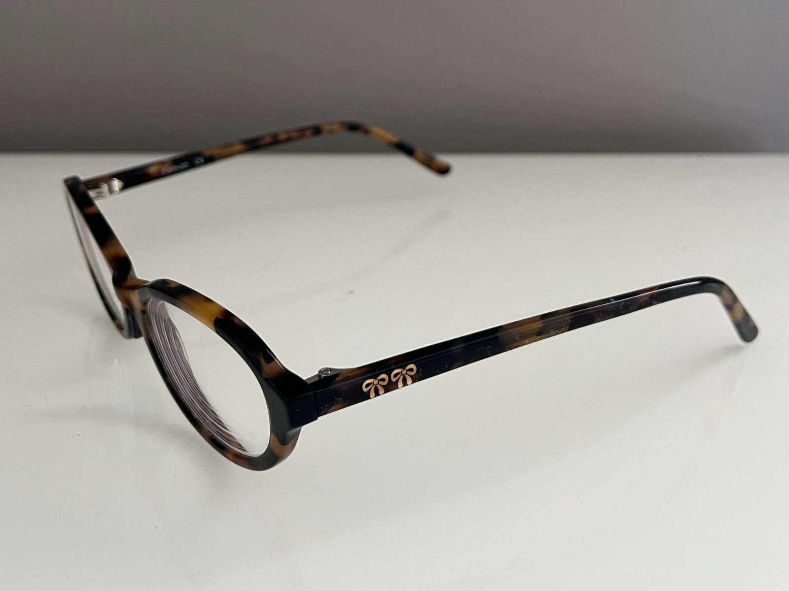 This photo shows a pair of tortoiseshell glasses on a white surface, taken from a diagonal side angle. The frame is slim with round lenses and thin arms, so the tortoiseshell pattern is clear along the temples. The gold detail on the arm is small but still visible. The angle shows the long shape of the glasses and their curve, while the shadow on the surface gives them more depth. This view also helps to see the size of the arms compared to the frame.