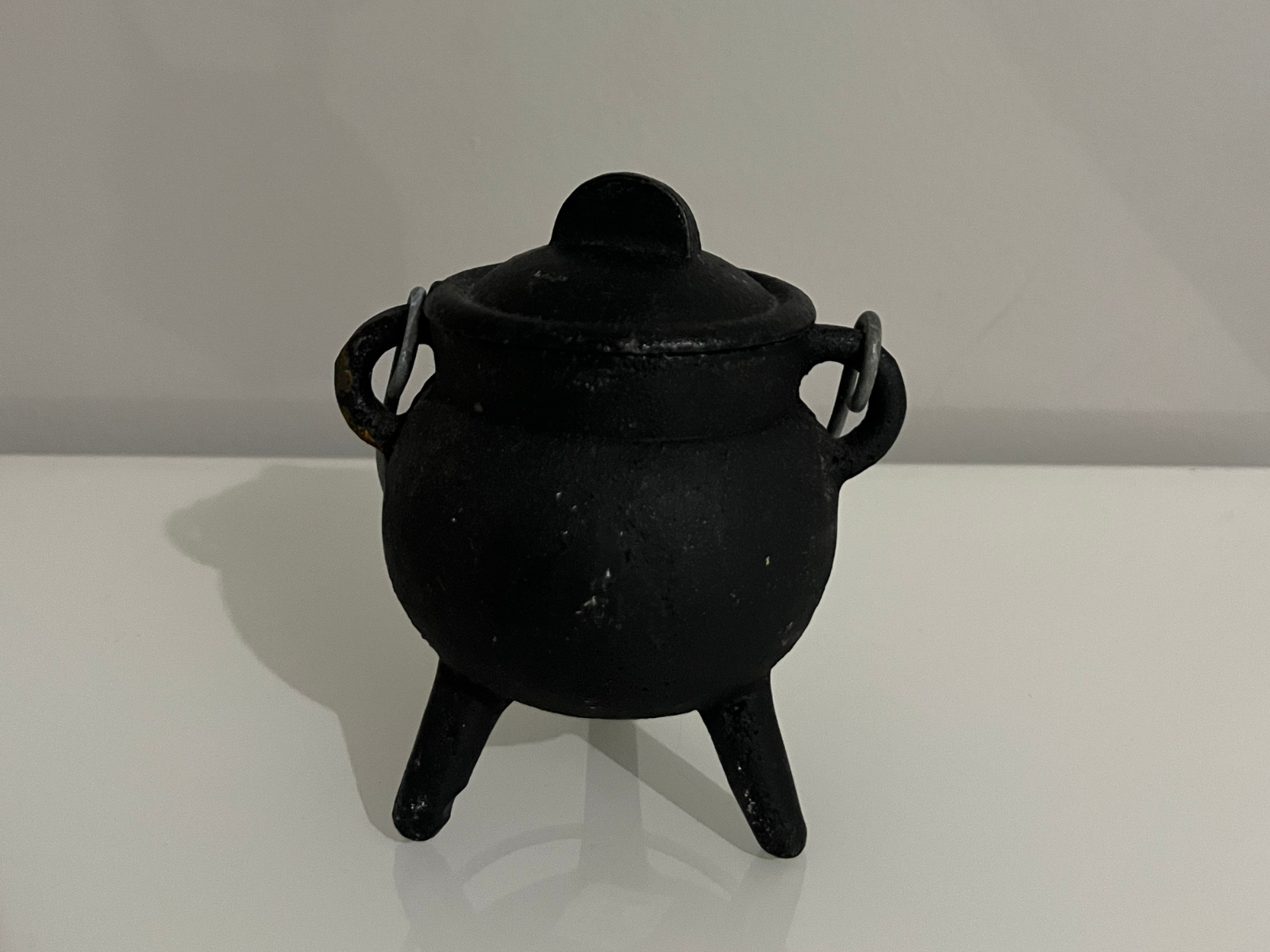 This is a reference photo of a small black cauldron, photographed from the front at eye level. The image highlights the rounded body supported by three short legs, with two small side handles and a fitted lid on top. The matte surface shows subtle texture and imperfections, giving it a realistic, worn look. The lighting creates a faint shadow on the white surface beneath it, emphasizing the shape and volume of the cauldron. This angle works well as a modeling reference since it clearly displays the proportions and overall silhouette.