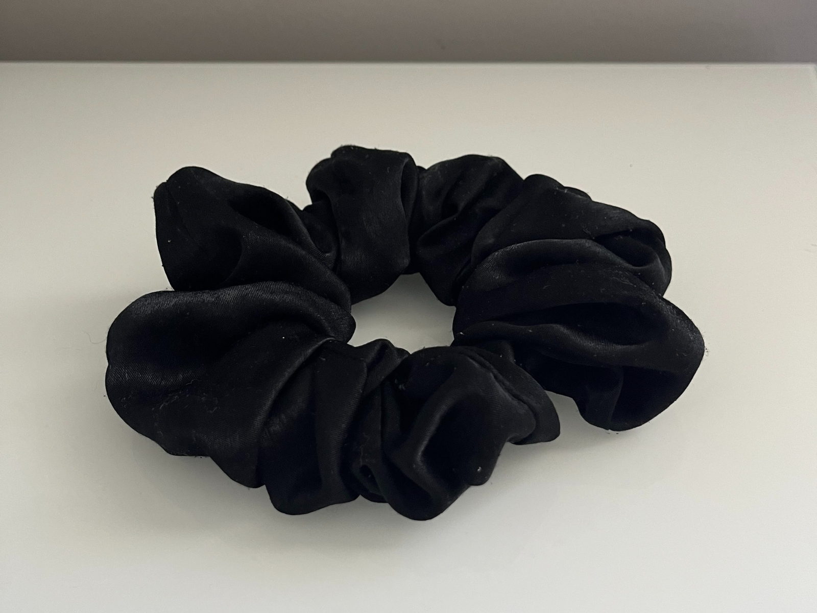 This is a photo of a black scrunchie, taken from the top view. The angle shows the round shape clearly and also the folds and texture of the fabric. The light makes a soft shine on the material, which gives it more depth and movement.