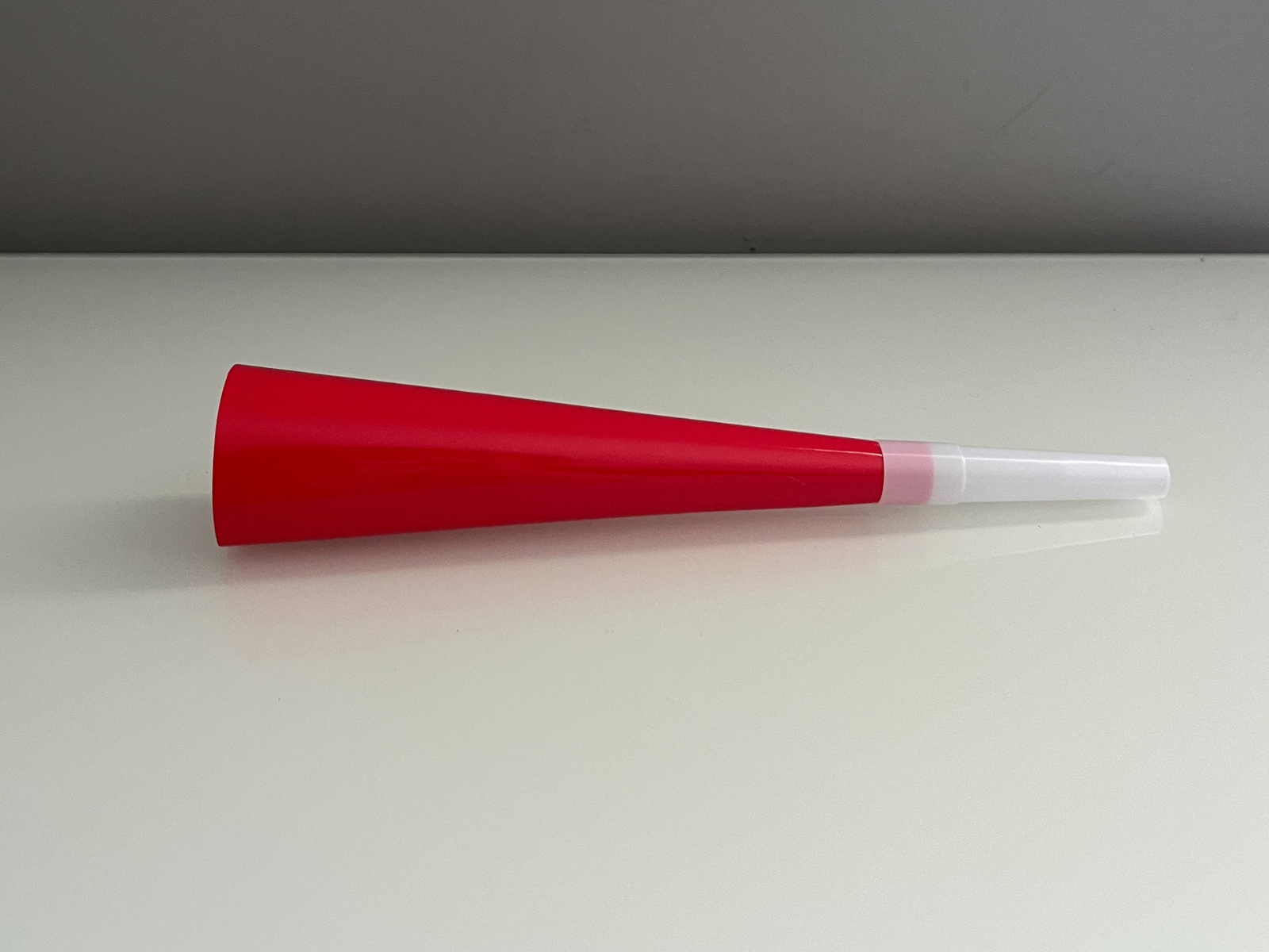 This is a photo of a red and white plastic horn, placed flat on a surface. The angle shows the full length and the cone shape, getting smaller toward the mouthpiece. The shiny plastic reflects the light, which makes the surface look smooth and the colors bright. There is also a small reflection under the horn that gives the image more depth.